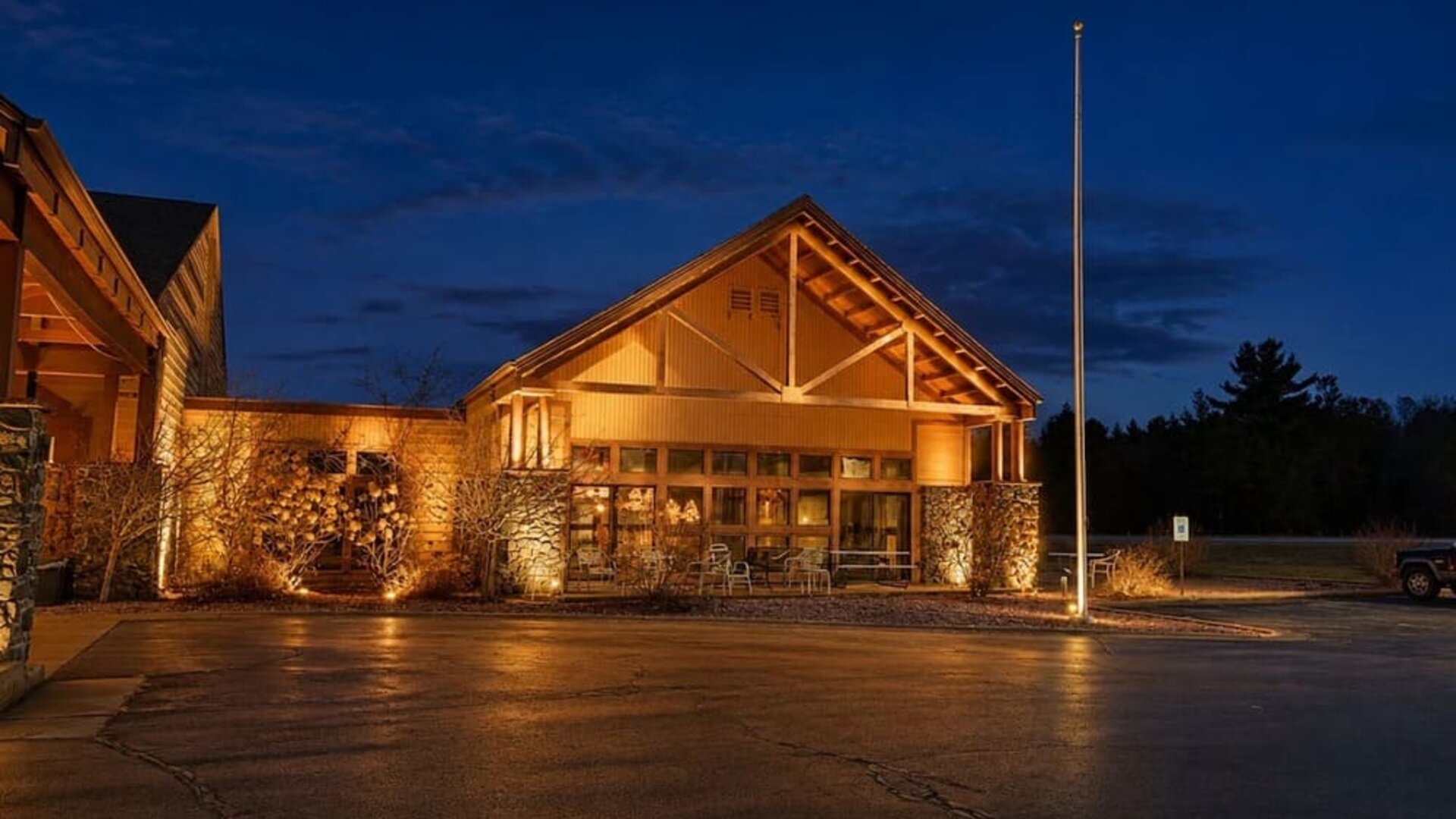 Lodge-style hotel exterior illuminated at night, featuring timber and stone architectural details, a peaked roof, large windows and outdoor seating near the entrance.