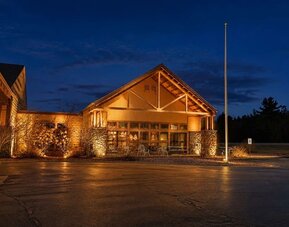 Lodge-style hotel exterior illuminated at night, featuring timber and stone architectural details, a peaked roof, large windows and outdoor seating near the entrance.