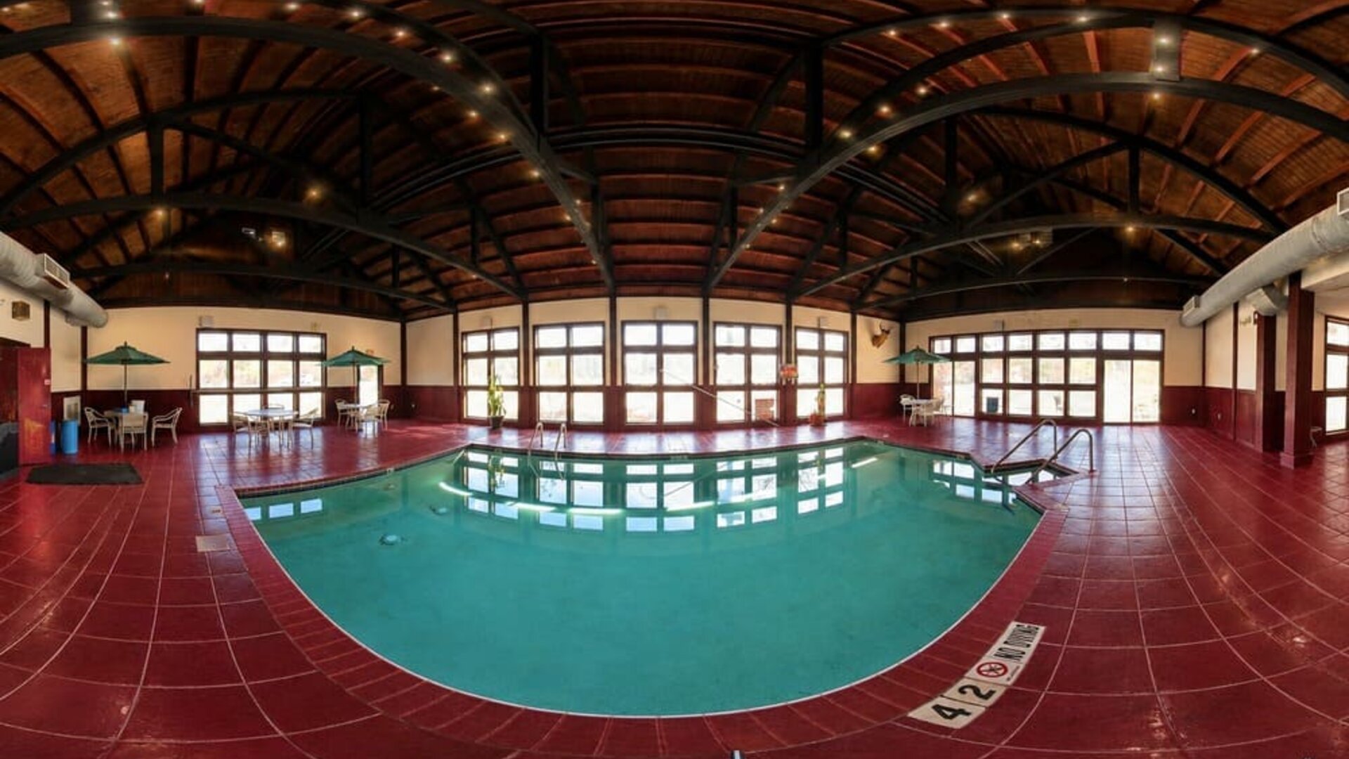 Spacious indoor hotel swimming pool surrounded by deep red tile flooring beneath a high, curved wood-paneled ceiling supported by dark exposed beams. Large windows and glass doors fill the pool area with natural light, while small seating areas with table