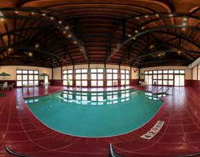 Spacious indoor hotel swimming pool surrounded by deep red tile flooring beneath a high, curved wood-paneled ceiling supported by dark exposed beams. Large windows and glass doors fill the pool area with natural light, while small seating areas with table
