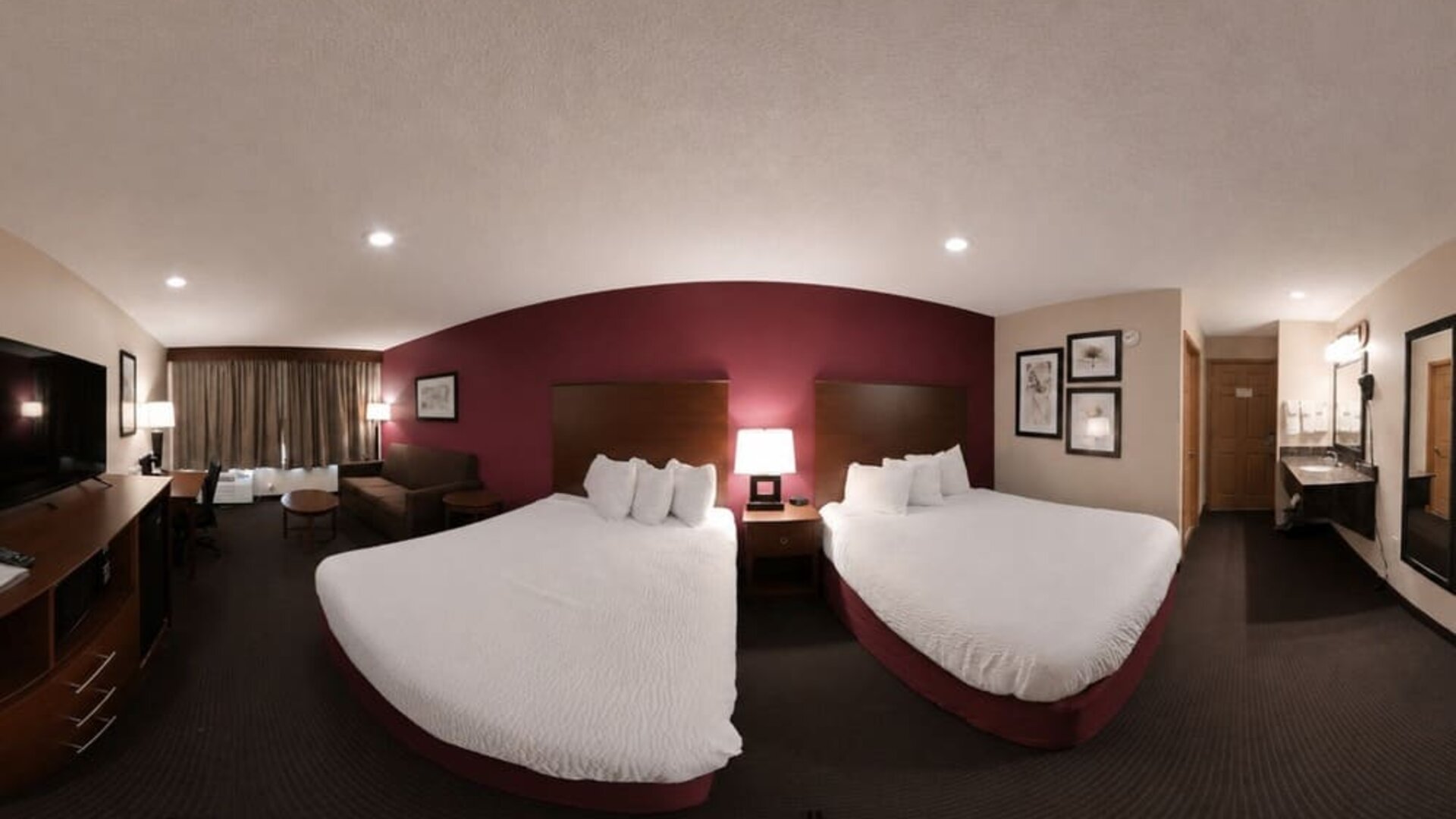 Comfortable hotel guest room featuring two beds with crisp white bedding and dark wooden headboards against a burgundy accent wall. The room includes a flat-screen TV, desk and chair, small seating area, bedside table with lamps, and curtained window. A v