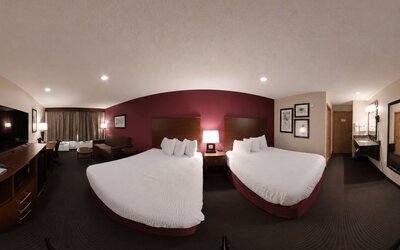 Comfortable hotel guest room featuring two beds with crisp white bedding and dark wooden headboards against a burgundy accent wall. The room includes a flat-screen TV, desk and chair, small seating area, bedside table with lamps, and curtained window. A v