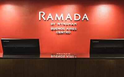 Hotel reception desk with two computer terminals and a red accent wall displaying the Ramada by Wyndham Buenos Aires Centro logo.