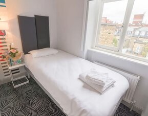 Single guest room with a white bed, dark upholstered headboard, bedside table, flowers and large window overlooking surrounding buildings at Victoria Inn.