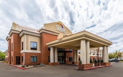 Exterior of Sleep Inn & Suites Stafford - Sugar Land, showing the brick-and-beige hotel building, covered entrance with columns, parking area, and landscaped planters.