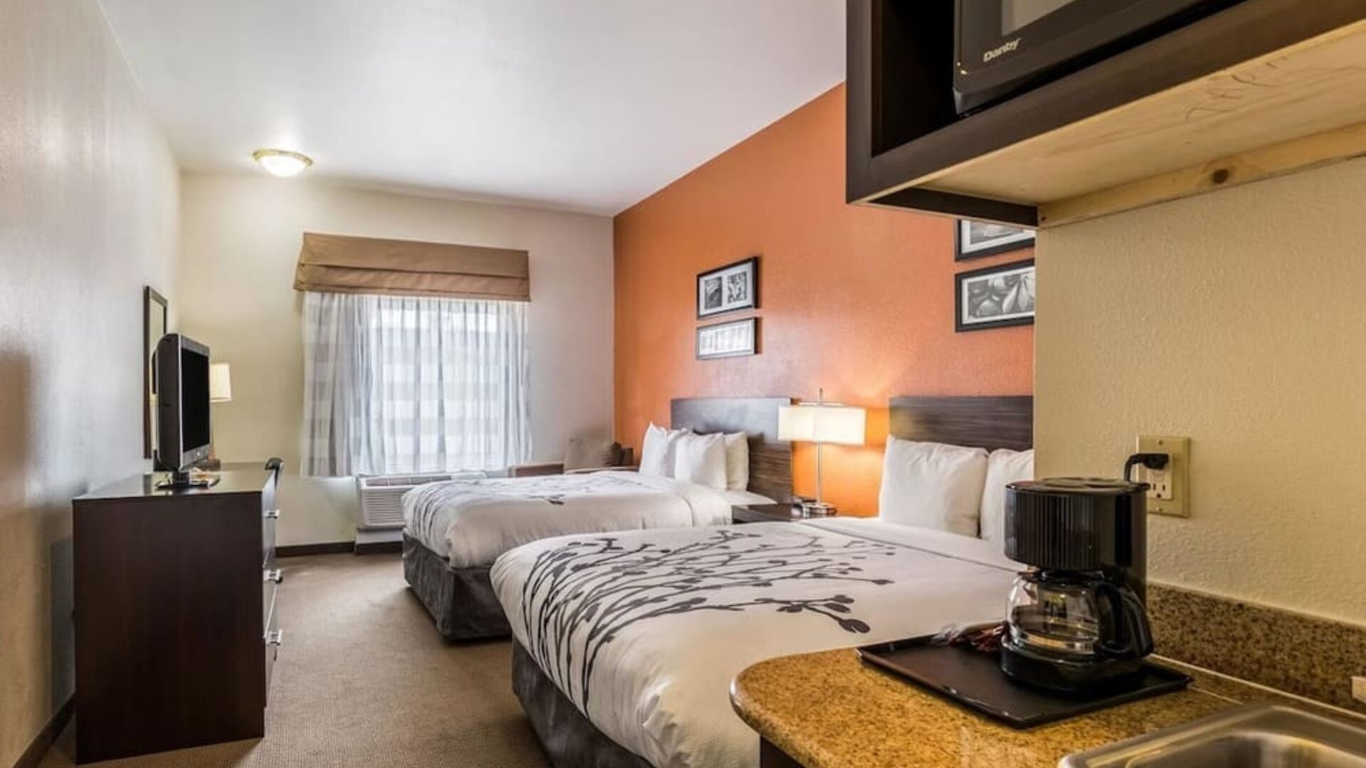 Double guest room with two beds, white bedding, orange accent wall, bedside lamps, television, microwave, refrigerator, coffee maker, and kitchenette counter at Sleep Inn & Suites Stafford - Sugar Land.