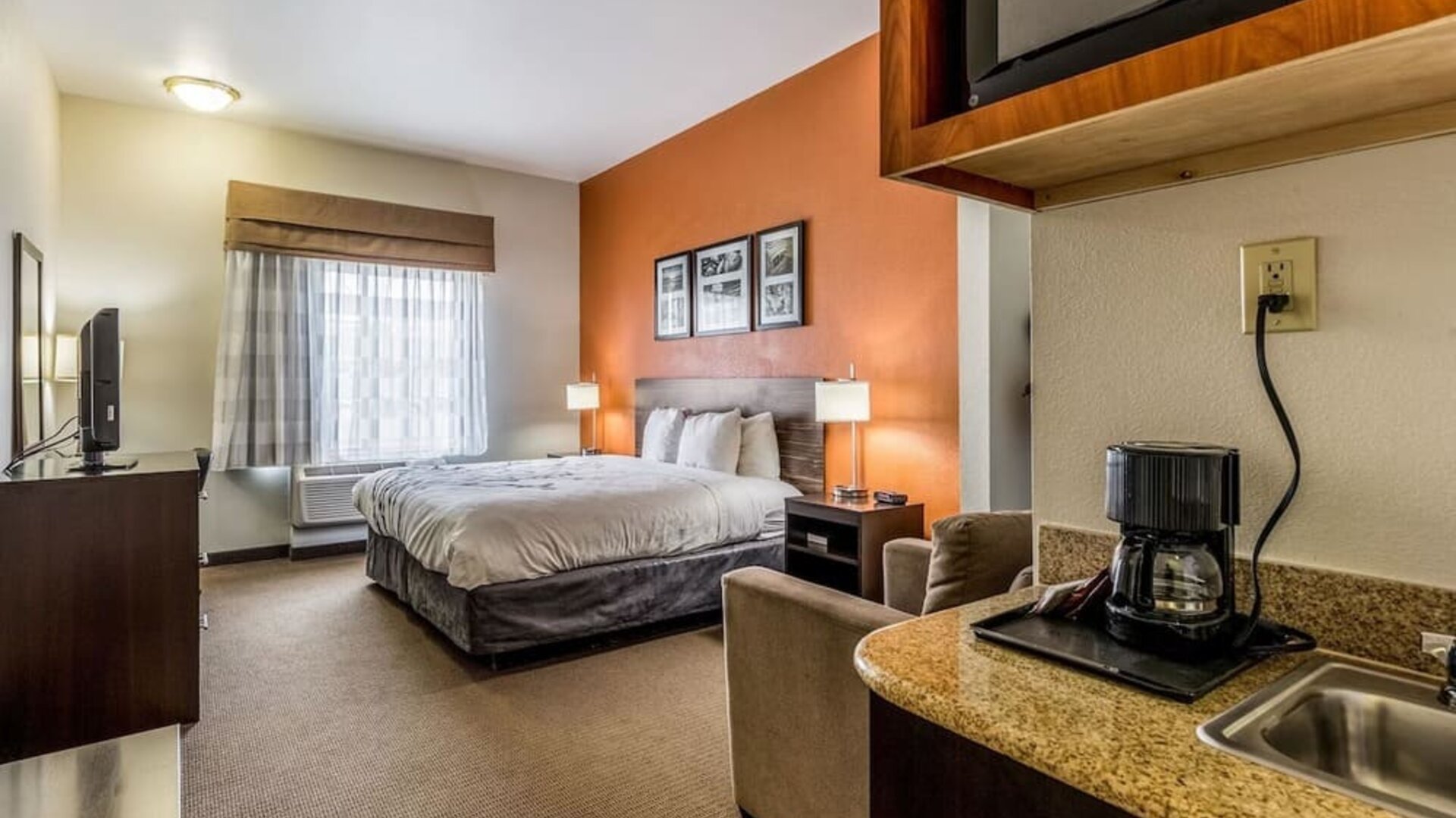 King guest room with a large bed, orange accent wall, framed artwork, bedside lamps, television, armchair, kitchenette counter, and coffee maker at Sleep Inn & Suites Stafford - Sugar Land.