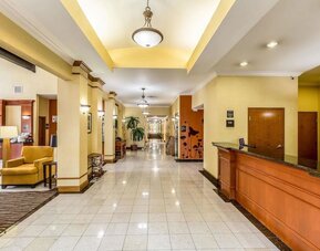 Hotel reception area with a long wooden front desk, yellow walls, seating area, tiled floor, and hallway leading to guest rooms at Sleep Inn & Suites Stafford - Sugar Land.
