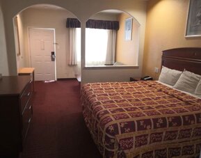 King guest room with a large patterned bed, dark wooden headboard, dresser, television, desk, mirror, and arched wall openings leading to the sitting area at Scottish Inn & Suites.