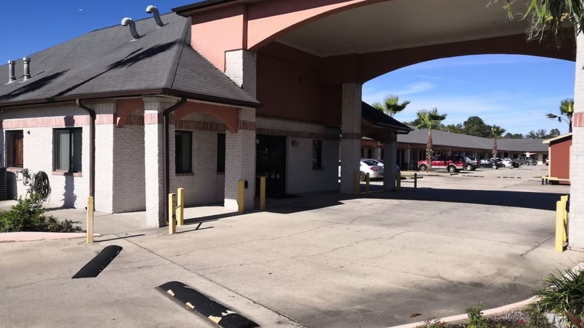 Exterior of Scottish Inn & Suites showing the low-rise hotel buildings, covered entrance with large arches, parking area, palm trees, and landscaped grounds.