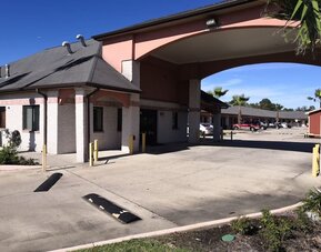 Exterior of Scottish Inn & Suites showing the low-rise hotel buildings, covered entrance with large arches, parking area, palm trees, and landscaped grounds.
