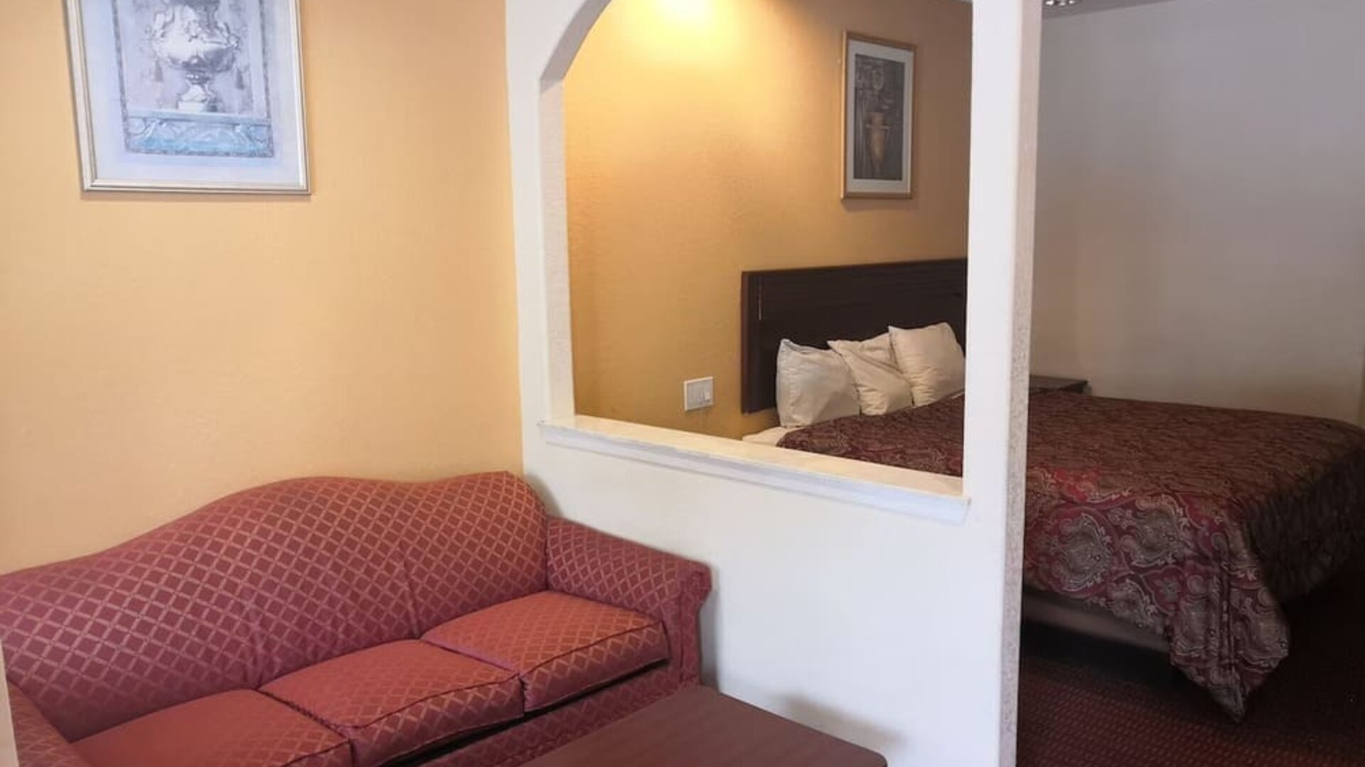 Guest room with a red patterned sofa and coffee table in the sitting area, with a king bed visible through two arched openings at Scottish Inn & Suites.