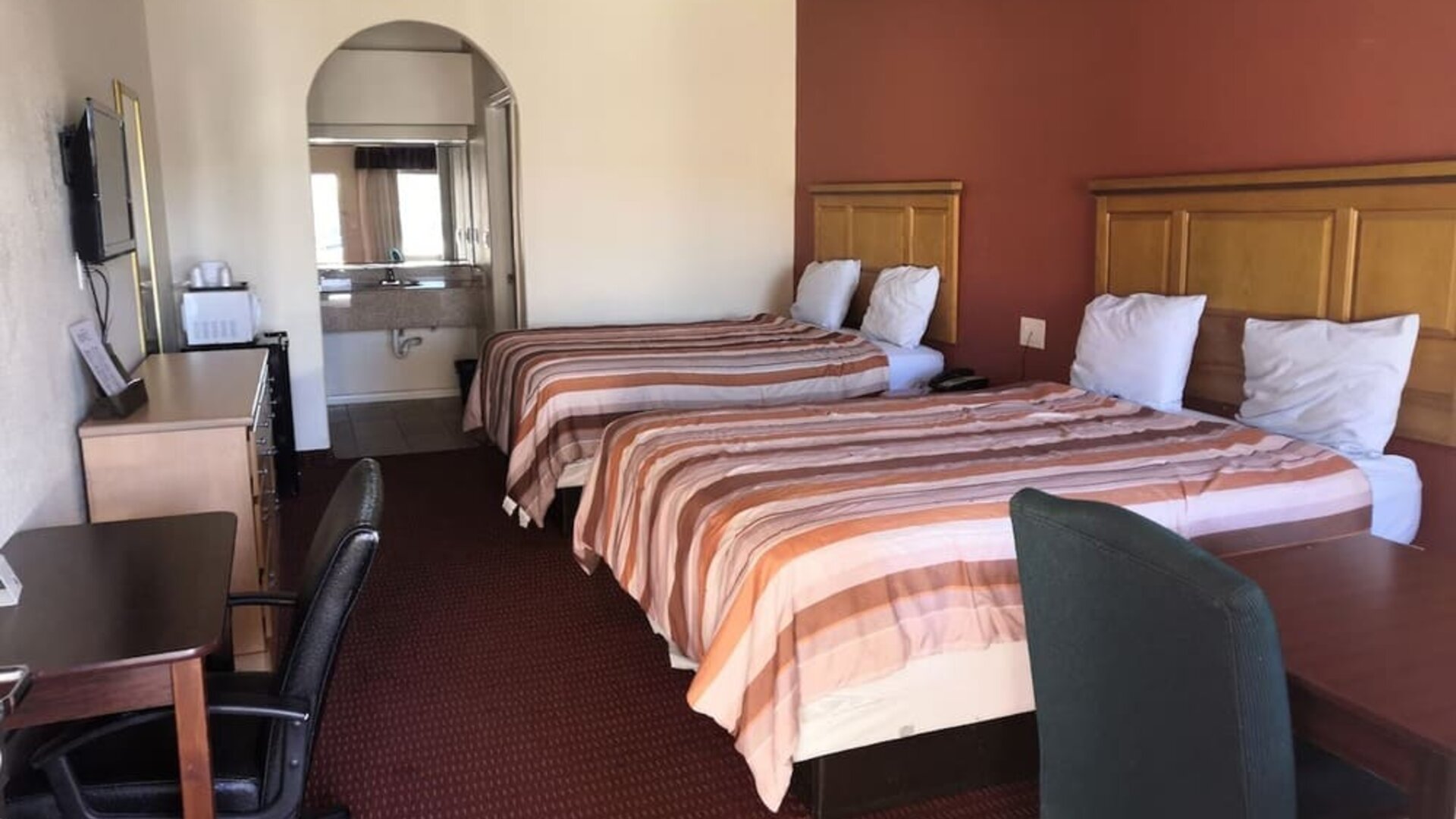 Guest room with two beds, striped orange and brown bedspreads, wooden headboards, desks and office chairs, television, and bathroom vanity visible through an arched opening at Scottish Inn & Suites.