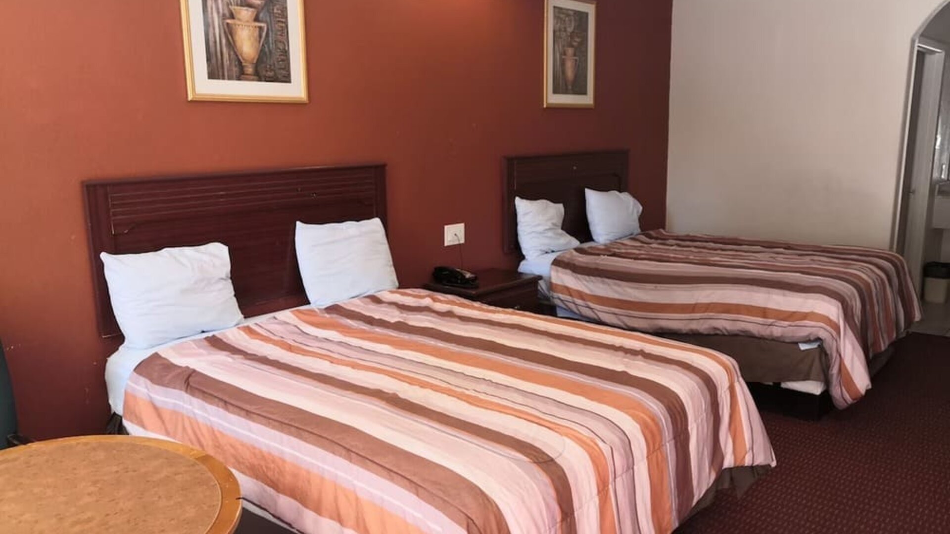 Guest room with two beds featuring striped orange, brown, and cream bedspreads, dark wooden headboards, bedside telephone, round table, and framed artwork at Scottish Inn & Suites.