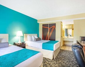 Guest room with two beds featuring white bedding and turquoise patterned runners, tufted headboards, bedside lamp, desk and chair, television, and bathroom vanity visible through an open doorway at Howard Johnson Express Inn - Rocky Hill.