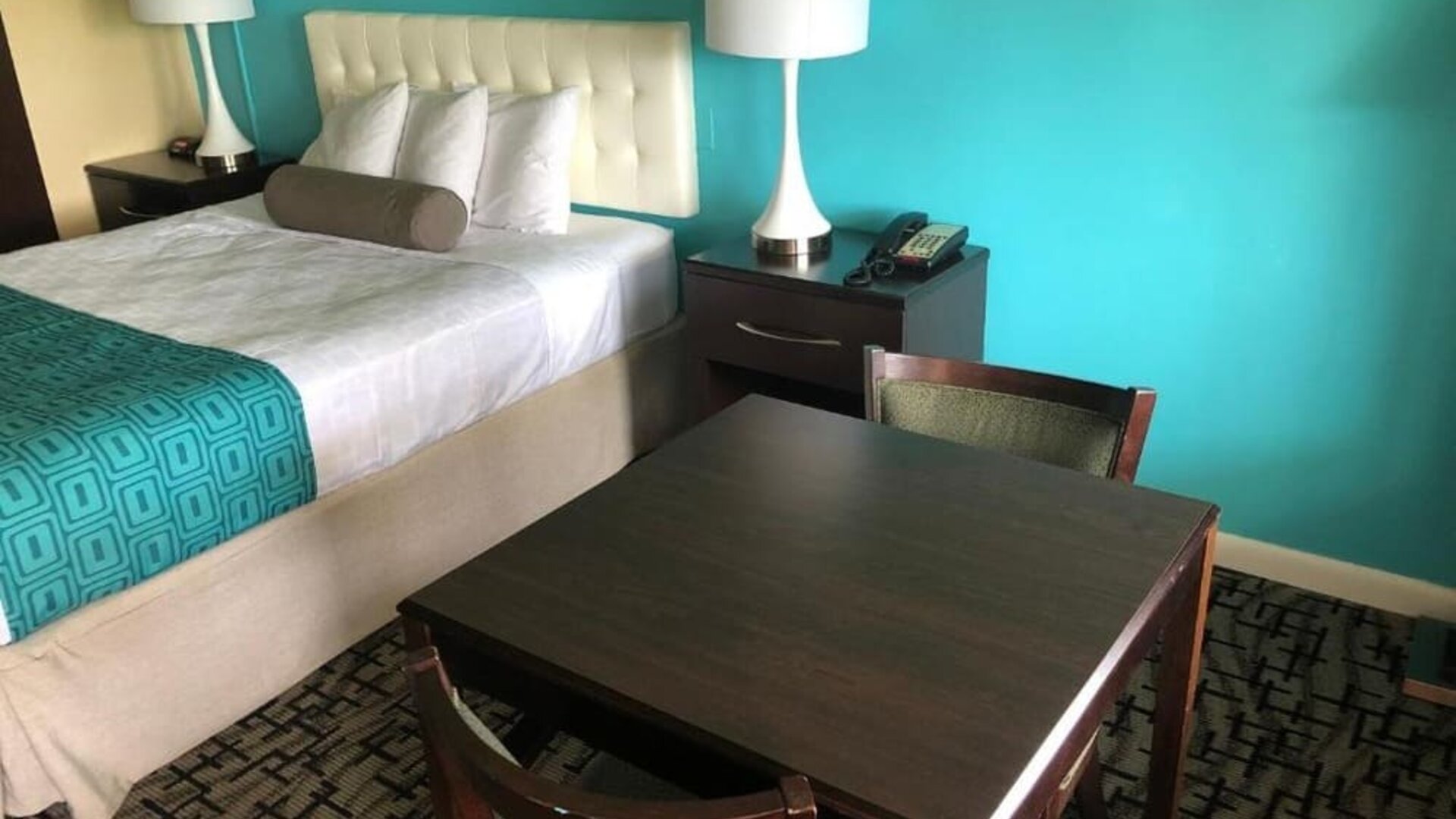 King guest room with a white tufted headboard, white bedding, turquoise patterned bed runner, dark bedside tables with lamps and telephone, and a dining table with chairs at Howard Johnson Express Inn - Rocky Hill.