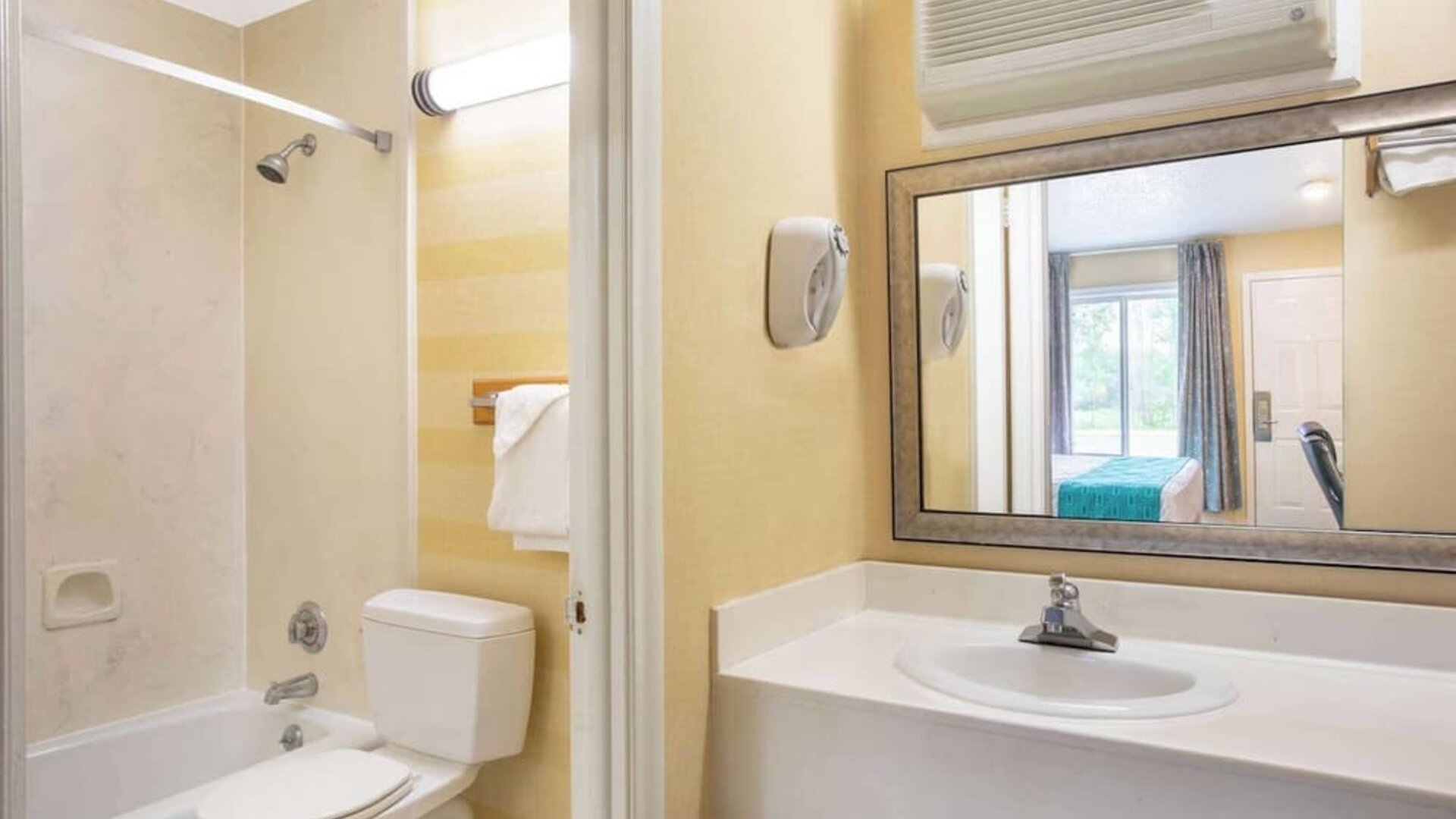 Bathroom with a bathtub and shower, toilet, white vanity with sink, large rectangular mirror, wall-mounted air-conditioning unit, and towel rack at Howard Johnson Express Inn - Rocky Hill.