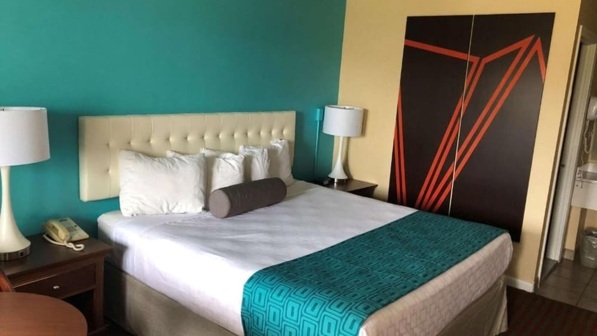 King guest room with white bedding, a tufted headboard, turquoise patterned bed runner, bedside tables with lamps and telephone, and large black and orange geometric wall artwork at Howard Johnson Express Inn - Rocky Hill.