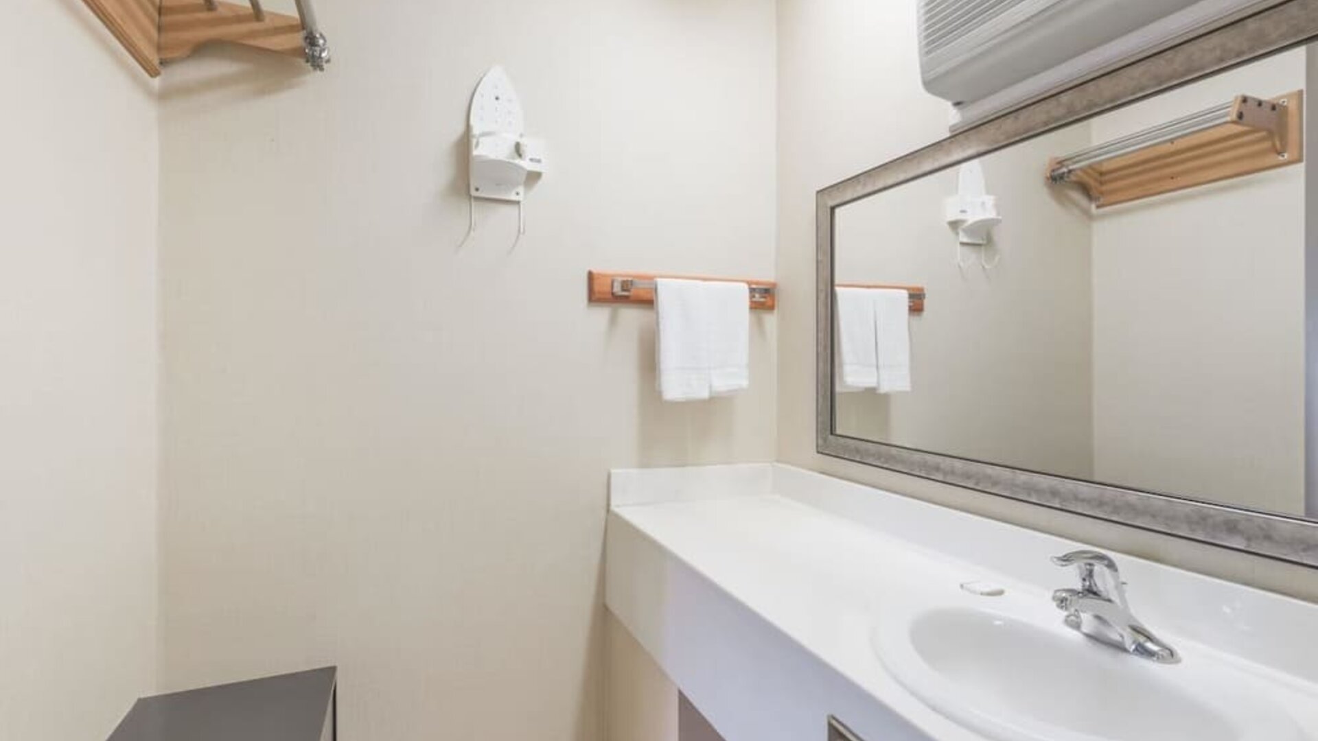 Bathroom vanity with a large rectangular mirror, white countertop and sink, towel rack with folded towels, wall-mounted hairdryer, and air-conditioning unit above the mirror at Howard Johnson Express Inn - Rocky Hill.