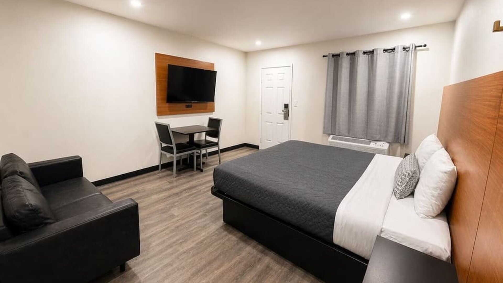 Bright contemporary king room featuring a large bed with gray and white bedding, oversized wood-paneled headboard, black loveseat, wall-mounted flat-screen TV, small dining table with two chairs, air-conditioning unit beneath gray curtains, recessed light
