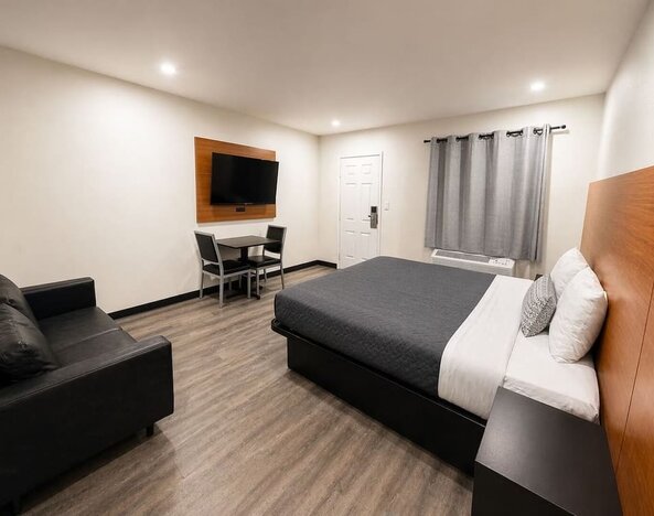 Bright contemporary king room featuring a large bed with gray and white bedding, oversized wood-paneled headboard, black loveseat, wall-mounted flat-screen TV, small dining table with two chairs, air-conditioning unit beneath gray curtains, recessed light