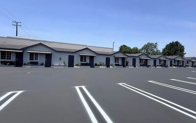 Daytime view of the Scenic Motel parking area with a clearly marked accessible parking space in the foreground and rows of single-story guest rooms with individual entrances, windows, small awnings, landscaped shrubs, and parking spaces extending into the