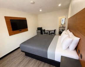 Bright modern guest room with a king-size bed dressed in gray and white bedding, a large wood-paneled headboard, wall-mounted flat-screen TV, small dining table with two chairs, microwave, mini refrigerator, wood-look flooring, and an open doorway leading
