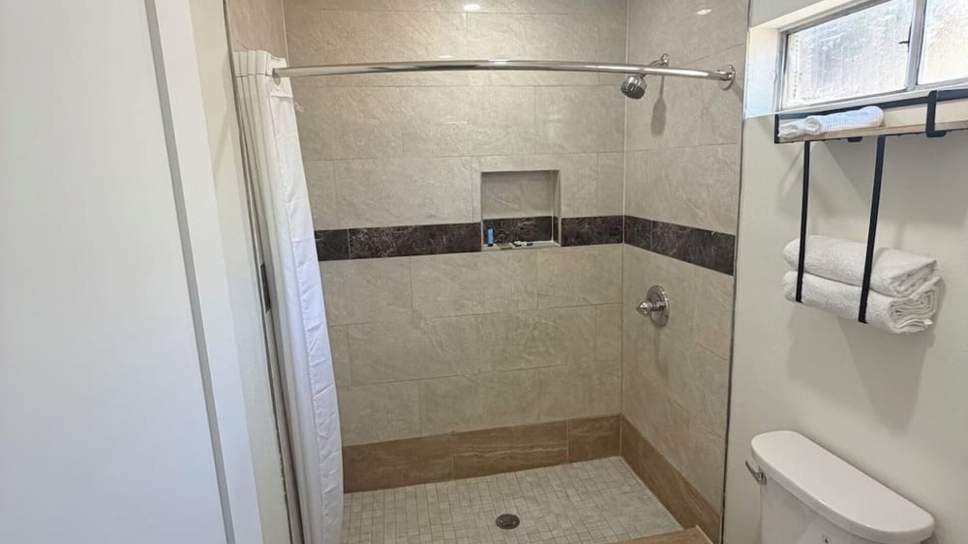 Modern private bathroom featuring a spacious tiled walk-in shower with a white curtain, built-in wall niche and showerhead, alongside a white toilet, wall-mounted towel rack with folded towels, frosted window, recessed lighting, and gray wood-look floorin