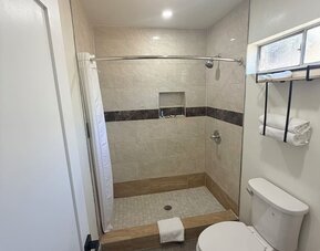 Modern private bathroom featuring a spacious tiled walk-in shower with a white curtain, built-in wall niche and showerhead, alongside a white toilet, wall-mounted towel rack with folded towels, frosted window, recessed lighting, and gray wood-look floorin