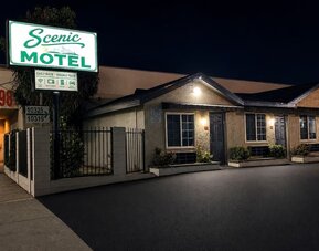 Nighttime exterior view of Scenic Motel with a large illuminated green-and-white roadside sign above the single-story property, where individual guest-room entrances, windows, exterior lights, landscaped planters, and the paved parking area are visible.