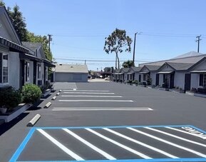 Wide exterior view of Scenic Motel showing a long row of single-story guest rooms with individual doors, windows, small covered awnings, and landscaped planters facing a large freshly paved parking area with clearly marked spaces under a bright blue sky.