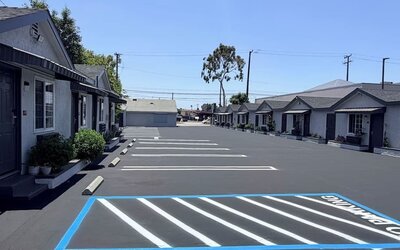 Wide exterior view of Scenic Motel showing a long row of single-story guest rooms with individual doors, windows, small covered awnings, and landscaped planters facing a large freshly paved parking area with clearly marked spaces under a bright blue sky.