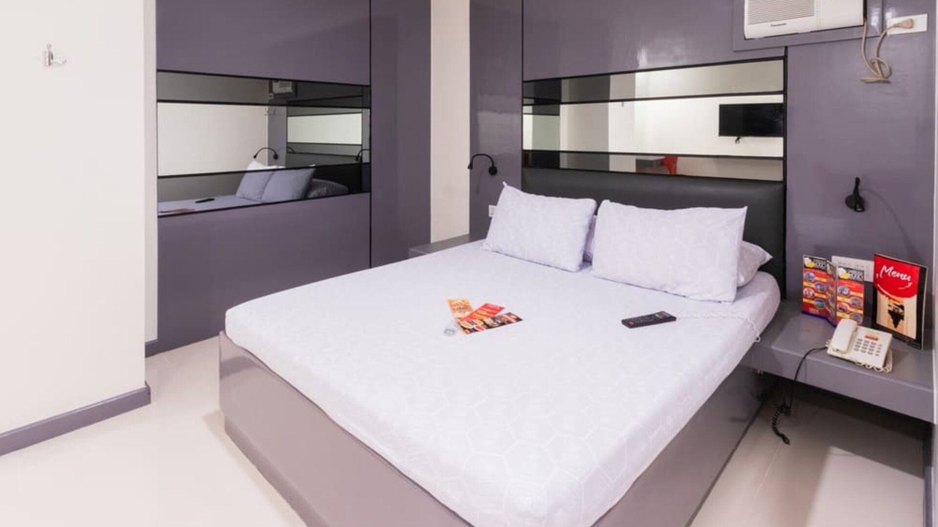 Modern double room with a white bed, purple-gray built-in headboard and mirrored panels, bedside shelves, reading lights, air-conditioning unit, telephone, and brochures at Hotel Sogo Guadalupe.