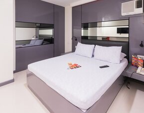 Modern double room with a white bed, purple-gray built-in headboard and mirrored panels, bedside shelves, reading lights, air-conditioning unit, telephone, and brochures at Hotel Sogo Guadalupe.