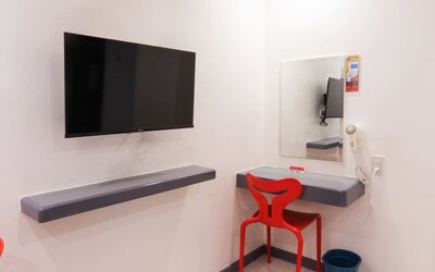 In-room workspace with a wall-mounted flat-screen TV, floating gray desk shelves, rectangular mirror, red chair, wall-mounted hairdryer, and waste bin at Hotel Sogo Guadalupe.