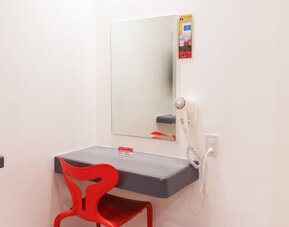 In-room workspace featuring a floating gray desk, rectangular wall mirror, bright red chair, wall-mounted hairdryer, and electrical outlet at Hotel Sogo Guadalupe.