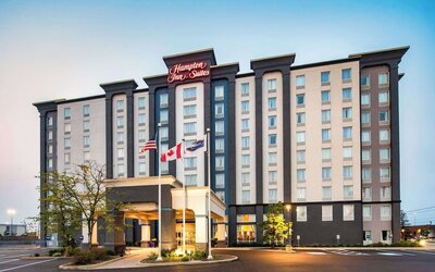 Exterior view of the multi-story Hampton Inn & Suites hotel with a covered entrance, landscaped grounds, parking area, and American, Canadian, and hotel flags displayed outside.
