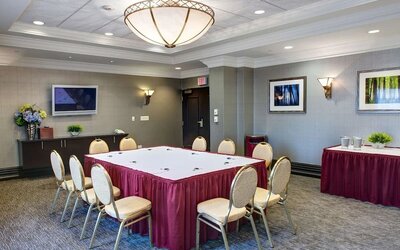 Hotel meeting room arranged with a U-shaped conference table, cushioned chairs, wall-mounted television, refreshment table, framed artwork, and overhead lighting.