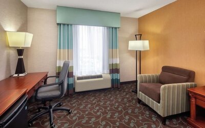 Hotel room work and relaxation area featuring a large wooden desk, ergonomic office chair, armchair, floor and desk lamps, mini refrigerator, and window with striped curtains.