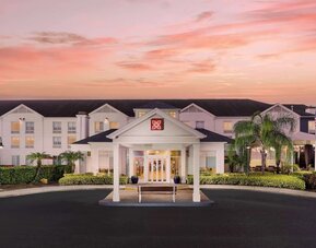 Front exterior of the hotel at sunset with a covered entrance, white three-story façade, palm trees, landscaped grounds and a pink-orange evening sky.