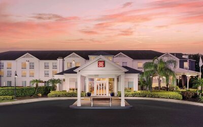 Front exterior of the hotel at sunset with a covered entrance, white three-story façade, palm trees, landscaped grounds and a pink-orange evening sky.