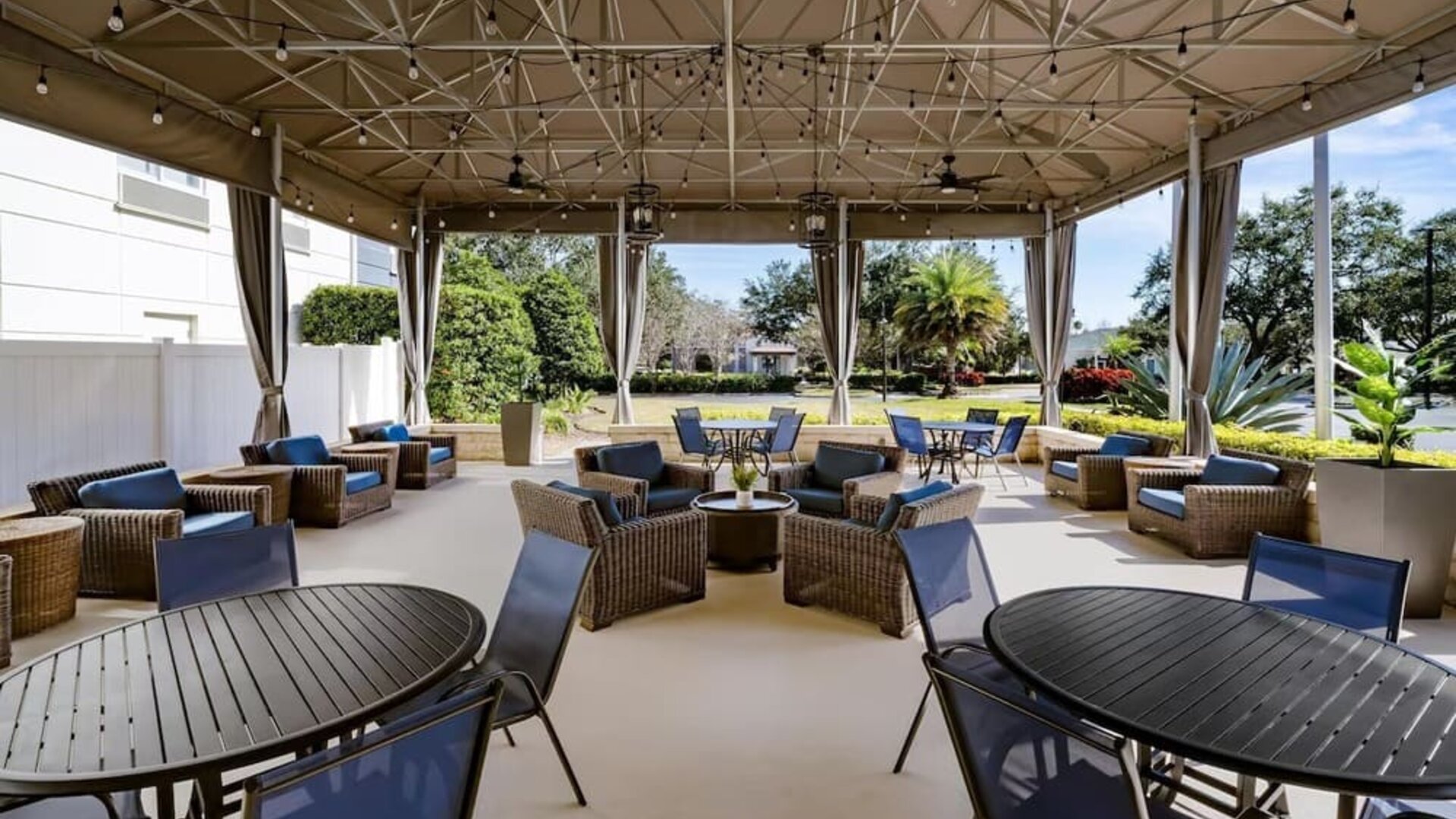 Covered hotel terrace with wicker lounge furniture, blue cushions, dining tables and chairs, string lights, ceiling fans and views of landscaped gardens.