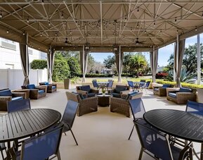 Covered hotel terrace with wicker lounge furniture, blue cushions, dining tables and chairs, string lights, ceiling fans and views of landscaped gardens.