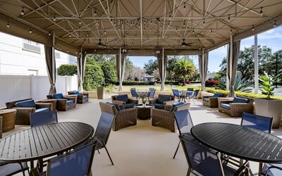 Covered hotel terrace with wicker lounge furniture, blue cushions, dining tables and chairs, string lights, ceiling fans and views of landscaped gardens.