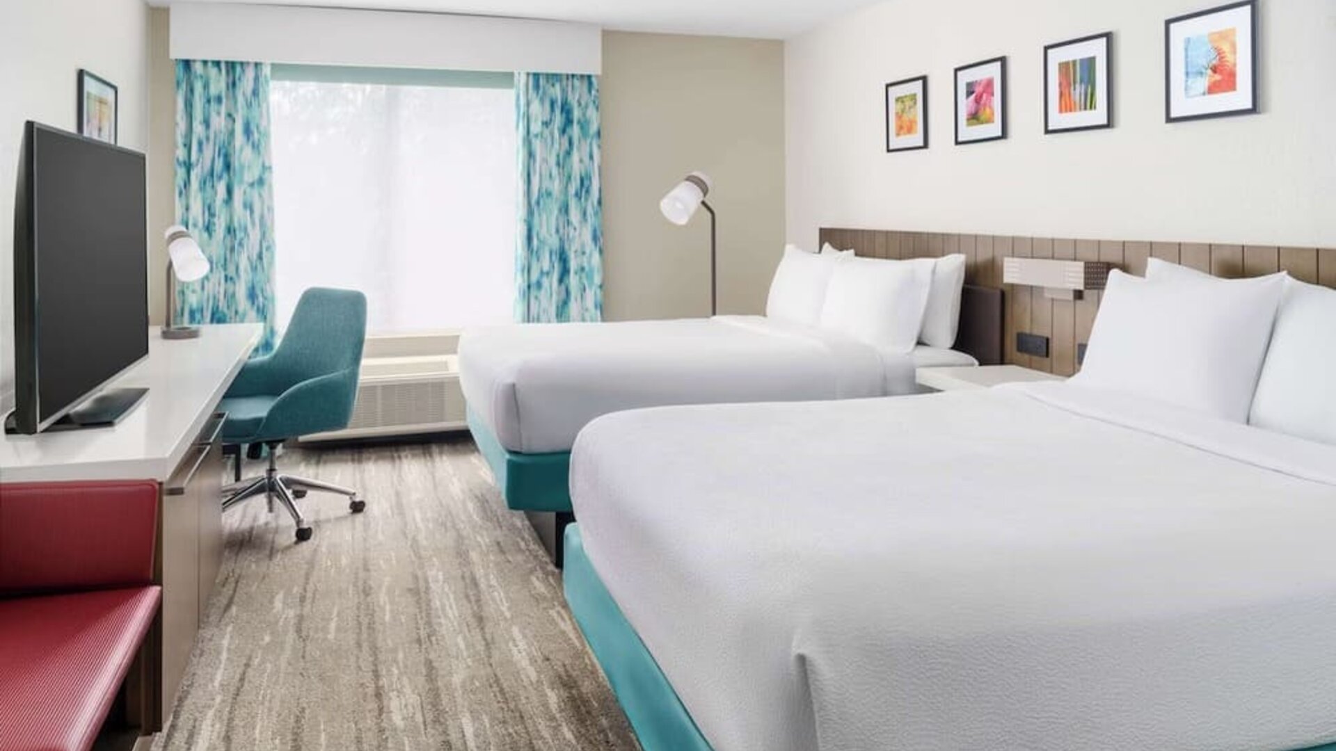 Bright modern hotel guest room with two white beds, turquoise bed bases, a large TV, work desk with teal chair, colorful artwork and patterned blue curtains.