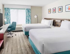 Bright modern hotel guest room with two white beds, turquoise bed bases, a large TV, work desk with teal chair, colorful artwork and patterned blue curtains.