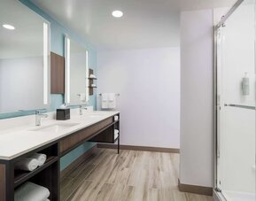Spacious modern hotel bathroom with a long double-sink vanity, illuminated mirrors, open towel storage and a glass-enclosed walk-in shower.