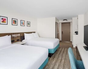 Spacious hotel guest room with two white beds on turquoise bases, colorful framed artwork, large flat-screen TV, teal desk chair and modern wood-look flooring.`