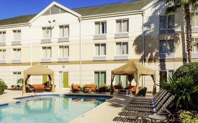 Outdoor hotel swimming pool surrounded by lounge chairs, shaded cabanas with orange seating, tropical landscaping and the hotel building.