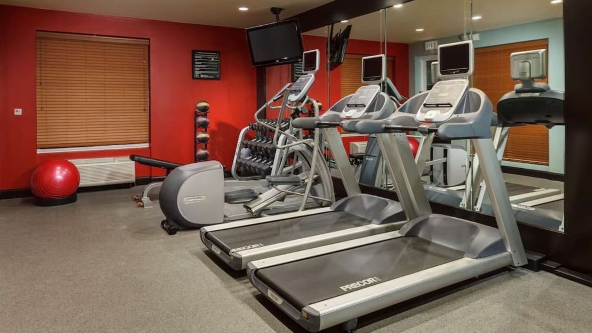 Hotel fitness center with two treadmills, elliptical machine, exercise bike, dumbbell rack, weight bench, stability ball, wall-mounted TV and mirrored wall.